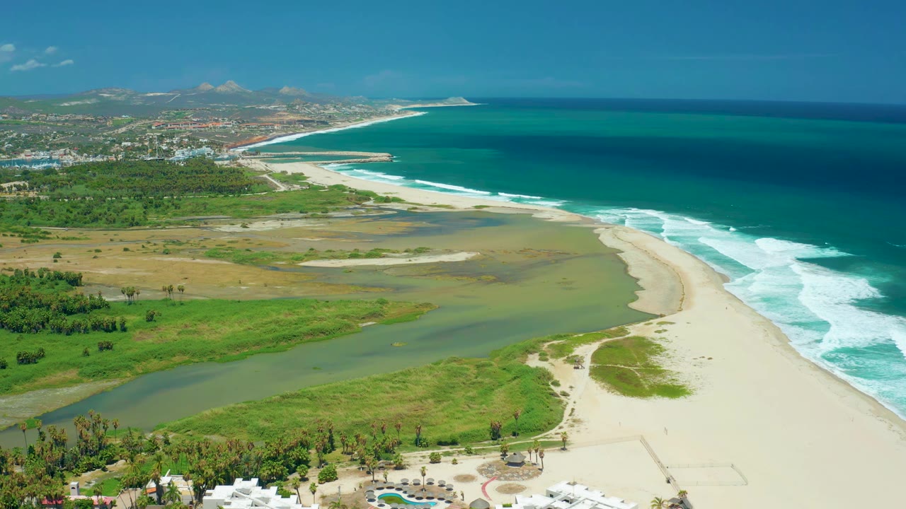 Wide aerial coast view with river water, sandbar, surf, vegetation, and hills.