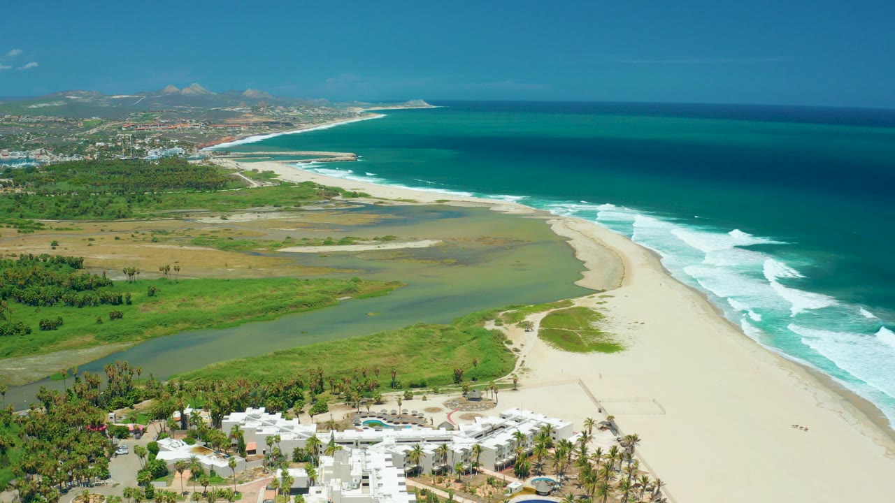 Opening aerial frame over San Jose del Cabo showing white resort roofs at the bottom, a green-blue river channel, pale sand, turquoise ocean, distant hills, and no visible people.