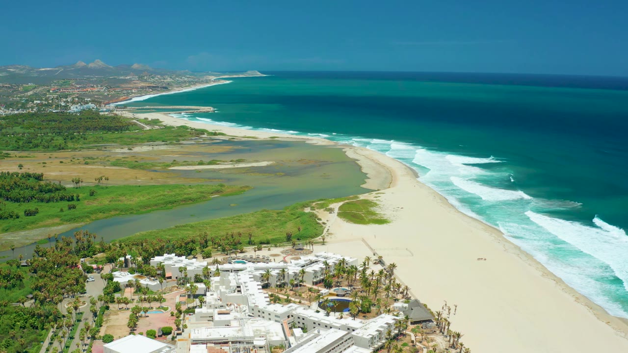 Aerial frame in San Jose del Cabo, Mexico with the sandbar and river channel centered below the beach curve, ocean waves breaking to the right, white buildings at the bottom edge, and no visible people.