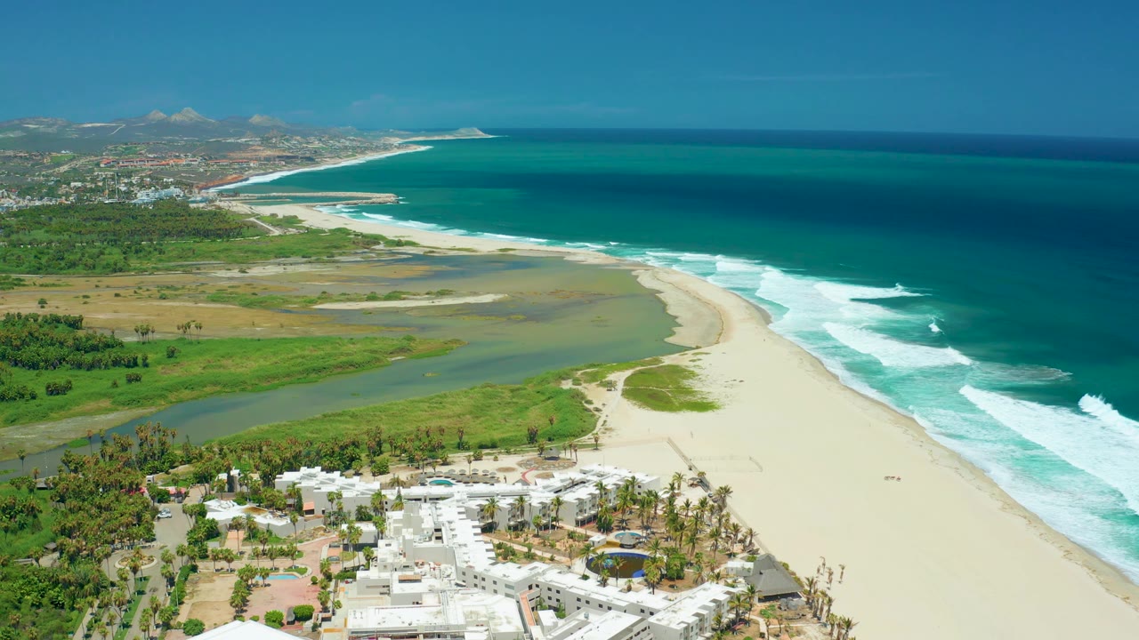 San Jose del Cabo aerial view showing the river lagoon cutting through green land toward the beach, white surf bands on the right, town development in the upper left, hills on the horizon, and no visible people.