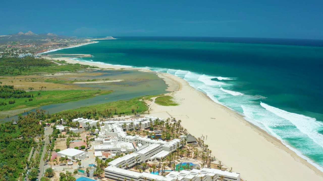 Drone view of curved beach, ocean surf, lagoon-like water, and resort-style buildings.