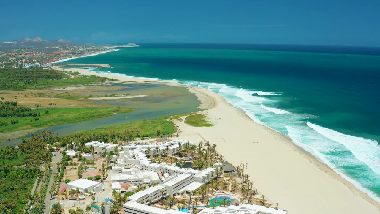 San Jose del Cabo aerial frame with white surf lines on the right, broad sand through the center, white coastal buildings at the lower edge, and no visible people.