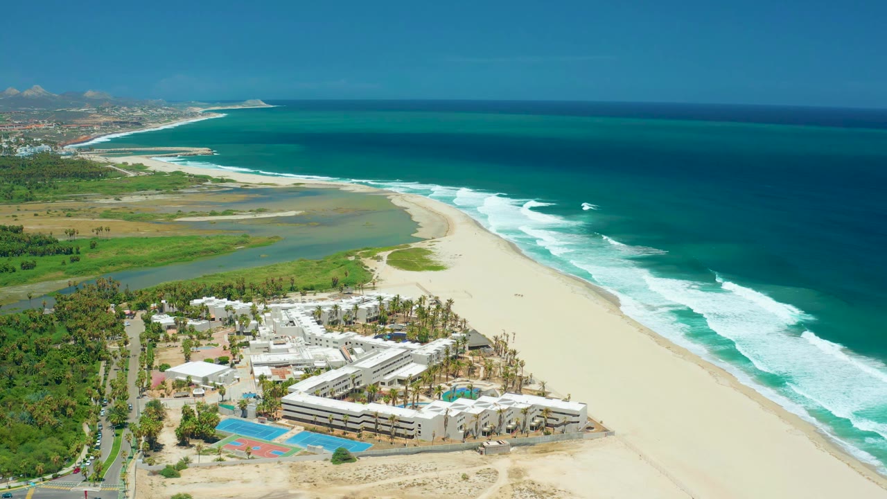 A high coastal view in San Jose del Cabo shows the lagoon curving behind the beach, surf breaking along the shore, hotel-style buildings and palms below, and no visible people.