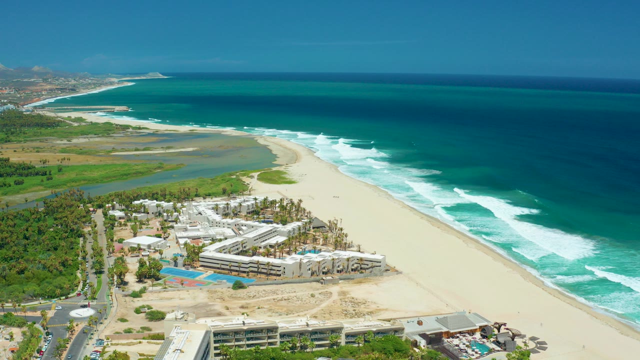 Aerial view of beach, turquoise ocean, resort buildings, pools, and coastal vegetation.