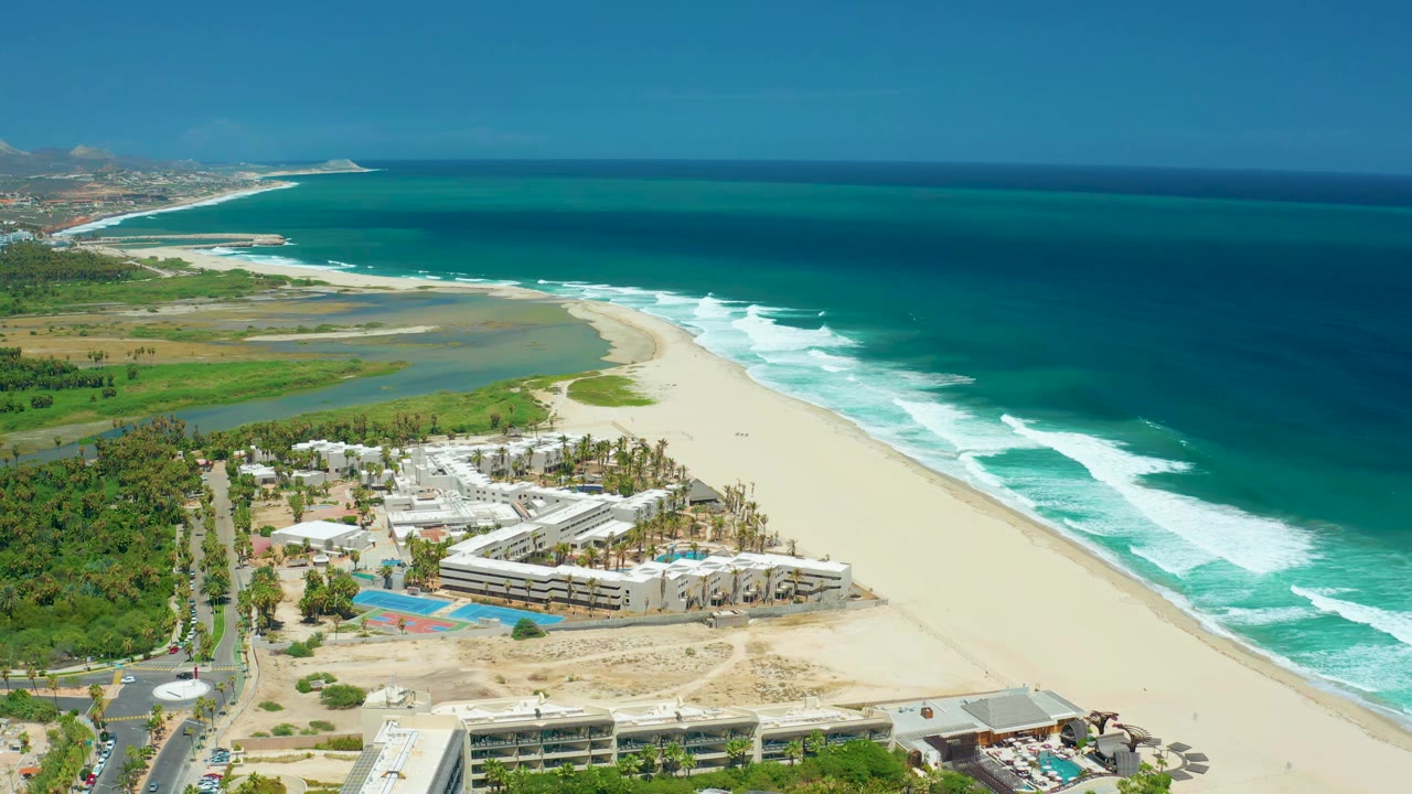 Aerial frame of San Jose Del Cabo coastline with a lagoon behind the beach, white waves along the shore, developed structures and pools at the lower edge, and no visible people.