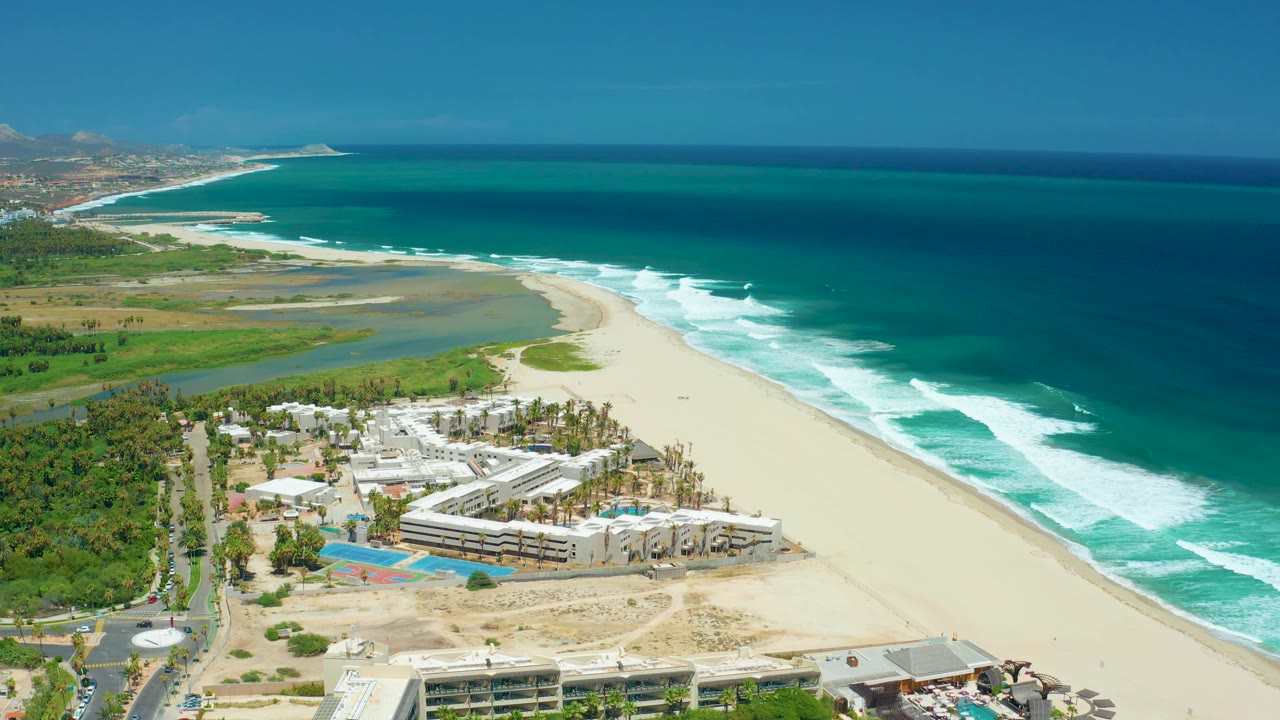 San Jose Del Cabo aerial view with foreground beachfront buildings, blue pool rectangles, pale sand, a curved lagoon edge, bright surf bands, deep blue water, and no visible people.