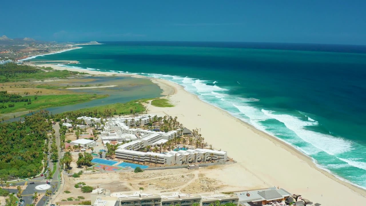 Aerial view over San Jose Del Cabo with a road and greenery at left, low beachfront structures and pools below, lagoon water behind the sand, rolling surf, and no visible people.