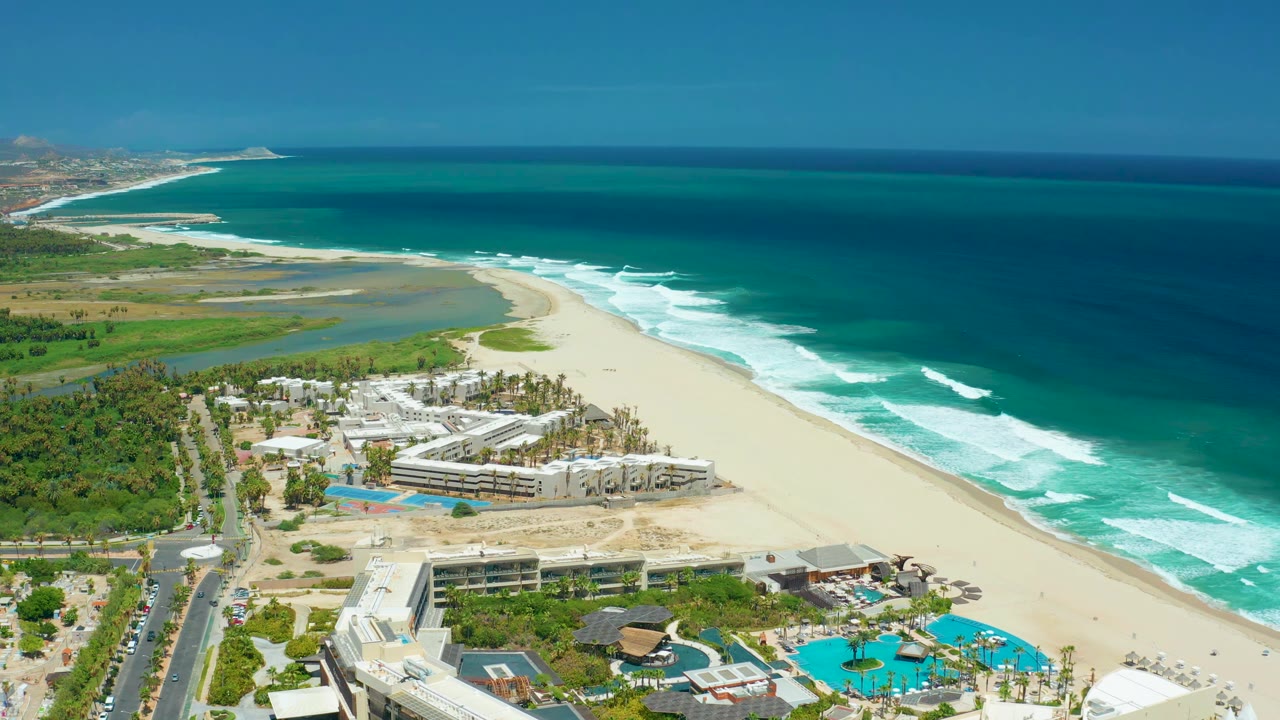 Aerial frame over San Jose del Cabo with resort buildings low in the view, pools near the foreground, a wide sand beach, rolling white surf, blue ocean, and no visible people.