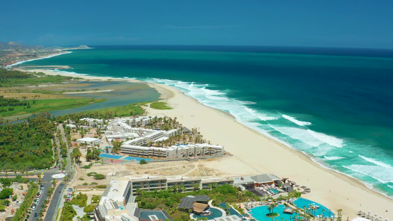 Aerial view of San Jose del Cabo coastline with beachfront hotel buildings, palm-lined grounds, road traffic areas, pale sand, white waves, blue ocean, and no visible people.