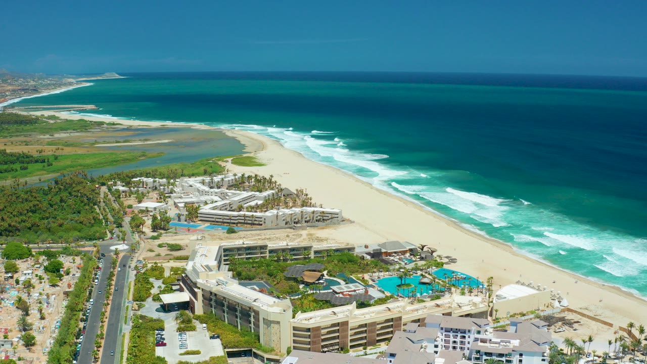 Aerial frame over San Jose Del Cabo, Baja California Sur, showing resort buildings and pools in the foreground, a sandy beach and lagoon behind them, and turquoise ocean with white surf beyond.