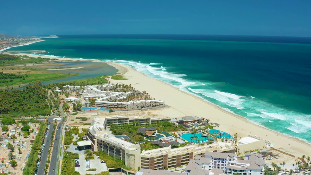 High aerial view in San Jose Del Cabo with beachfront resort blocks, blue pools, a road with cars at left, a pale sand beach, lagoon water, and ocean surf bands rolling toward shore.