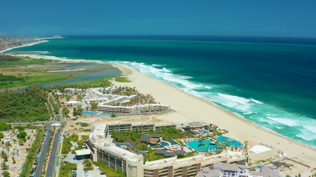 Aerial coastline view with resort rooftops, pool decks, sand, lagoon, surf, road, and cars.