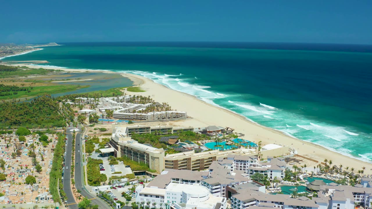 San Jose Del Cabo aerial frame showing the resort centered below the beach curve, white surf bands offshore, a road at left, and no visible people.