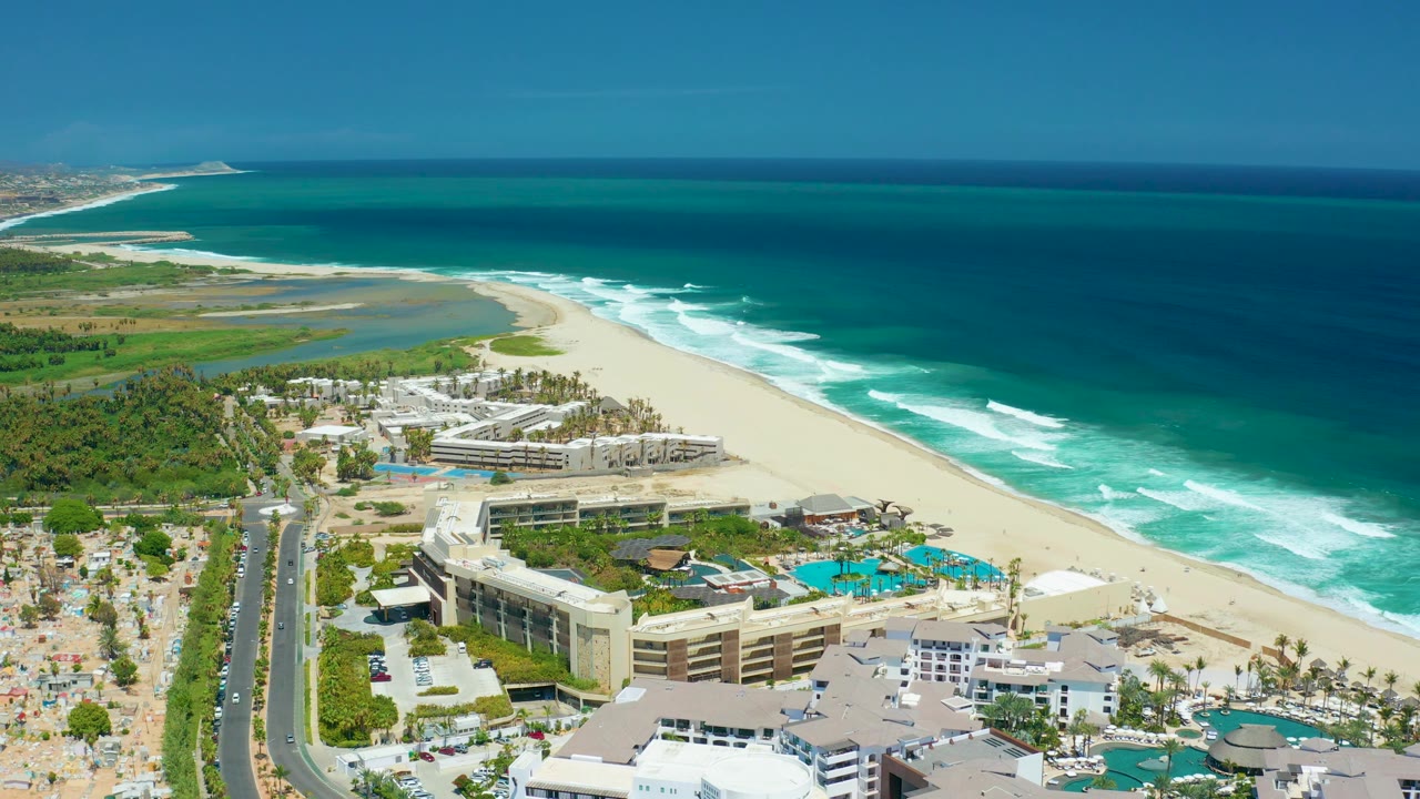 Mid-clip San Jose Del Cabo frame with resort roofs and pools in the foreground, a wide strip of sand and breaking waves in the middle, and no visible people.