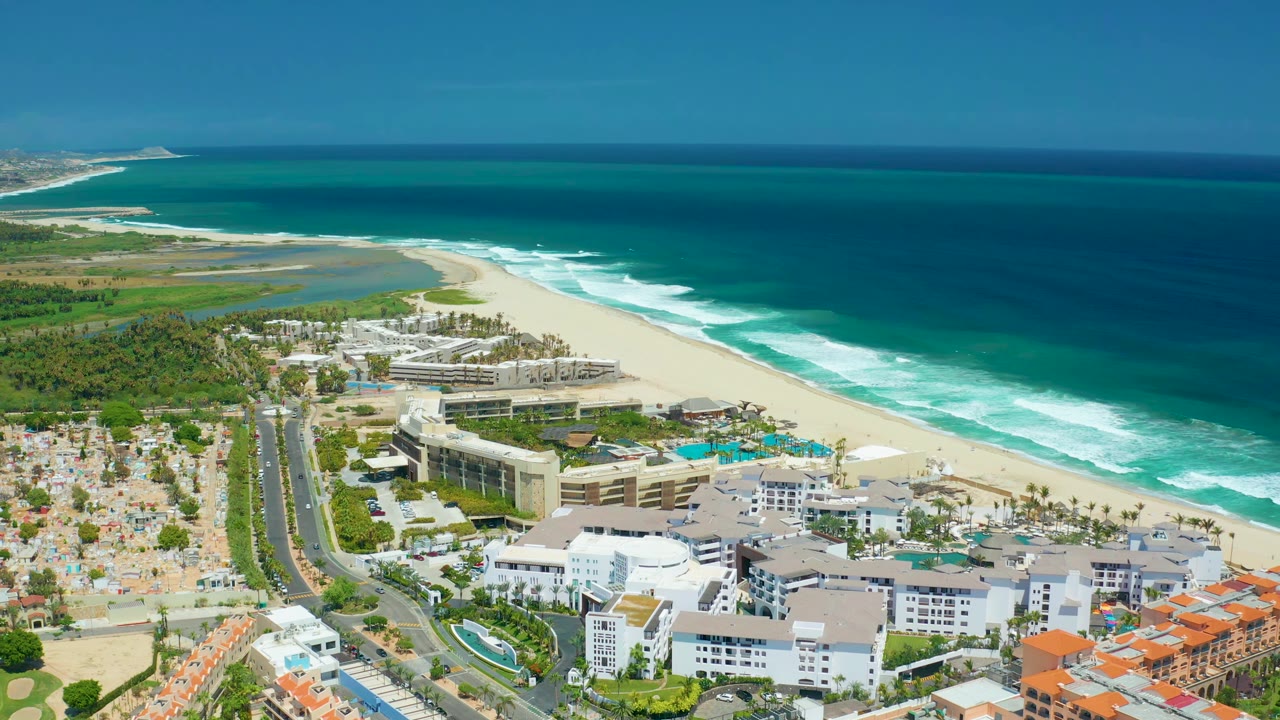 Aerial view in San Jose Del Cabo with white resort blocks and pools in the foreground, small vehicles on the road, and white surf lines moving along the beach, with no visible people.