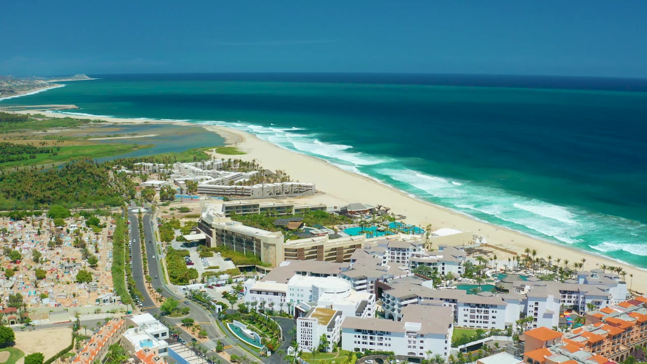San Jose Del Cabo aerial frame showing hotel-style buildings, pool decks, a road corridor, green coastal vegetation, a lagoon-like water area, sandy beach, and dark blue ocean, with no visible people.