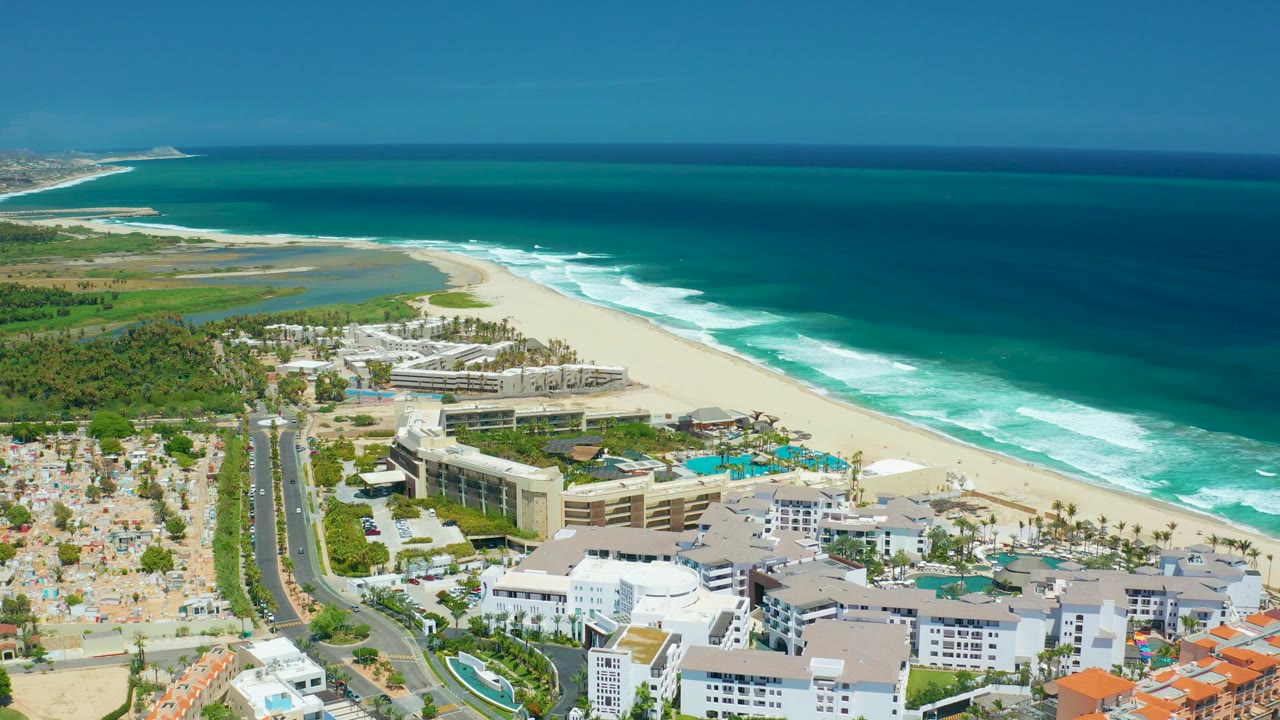 Wide aerial view over San Jose Del Cabo with the divided road and palms at left, resort buildings and pools below, and bright white waves breaking along the beach, with no visible people.