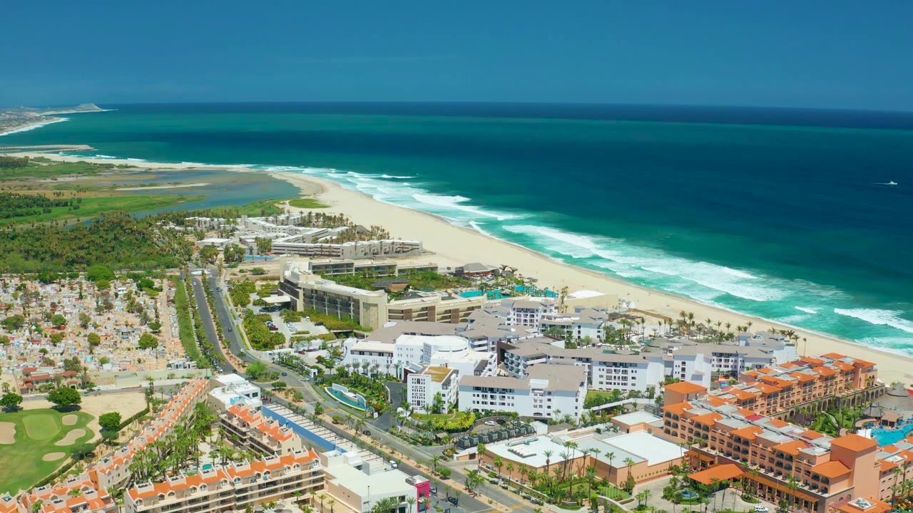 Aerial view one second into the San Jose Del Cabo clip, with surf bands running along the beach, lagoon water behind the sand, clustered buildings, roads, and no visible people.