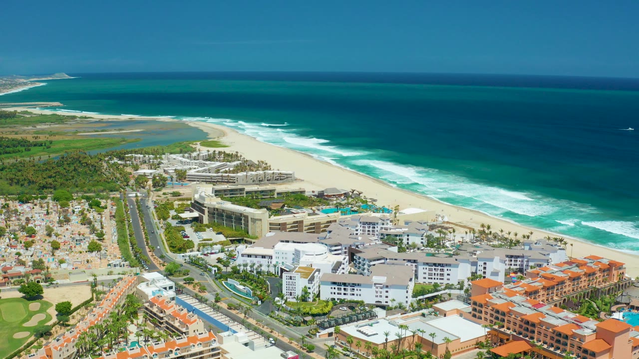 Mid-clip aerial frame over San Jose Del Cabo showing more open ocean across the top, white waves on the beach, lagoon water inland, shoreline buildings, roads, and no visible people.