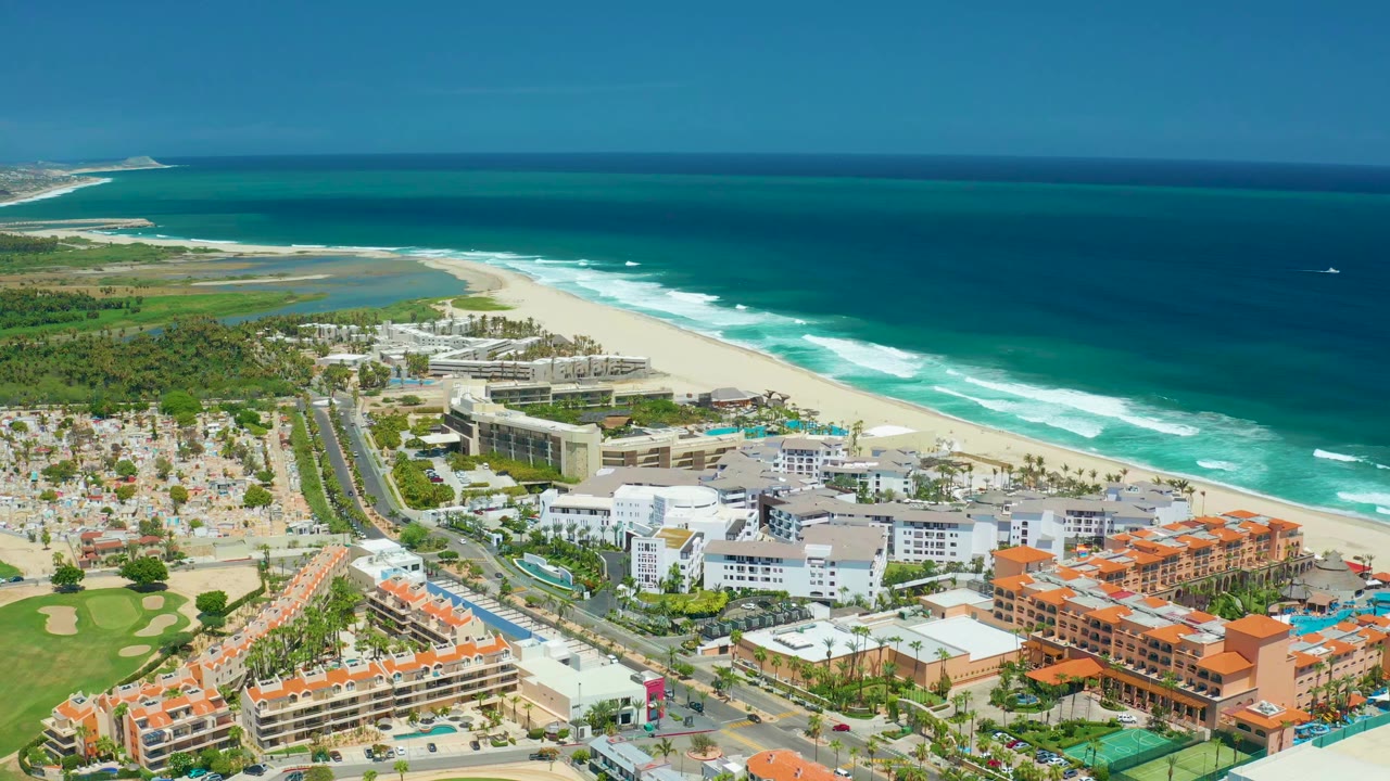Aerial view in San Jose Del Cabo with the coastal road and buildings lower in frame, bright beach and surf in the middle, and no visible people.