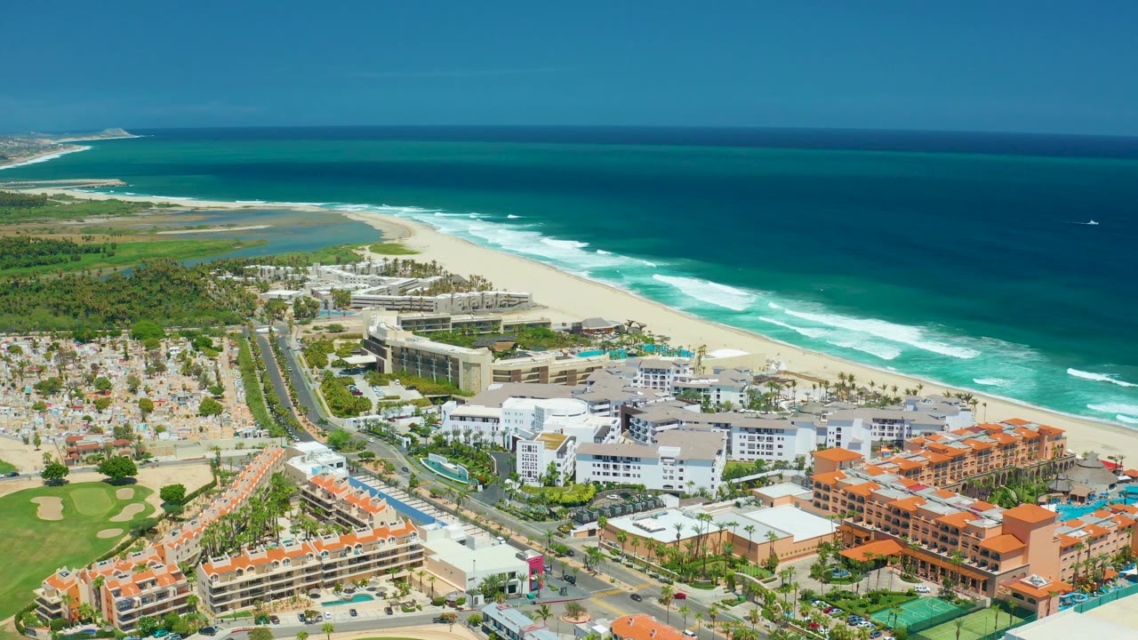 San Jose Del Cabo aerial frame showing resort-style beachfront buildings beside the road, a broad sand strip, layered surf, open ocean, and no visible people.