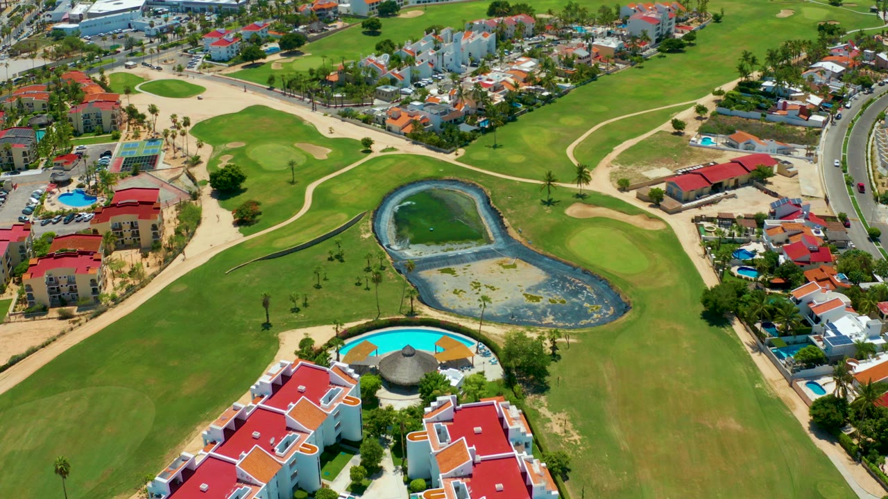 Aerial view over San Jose Del Cabo showing red-roofed homes and a bright pool in the foreground, a golf fairway and pond in the center, and no visible people.