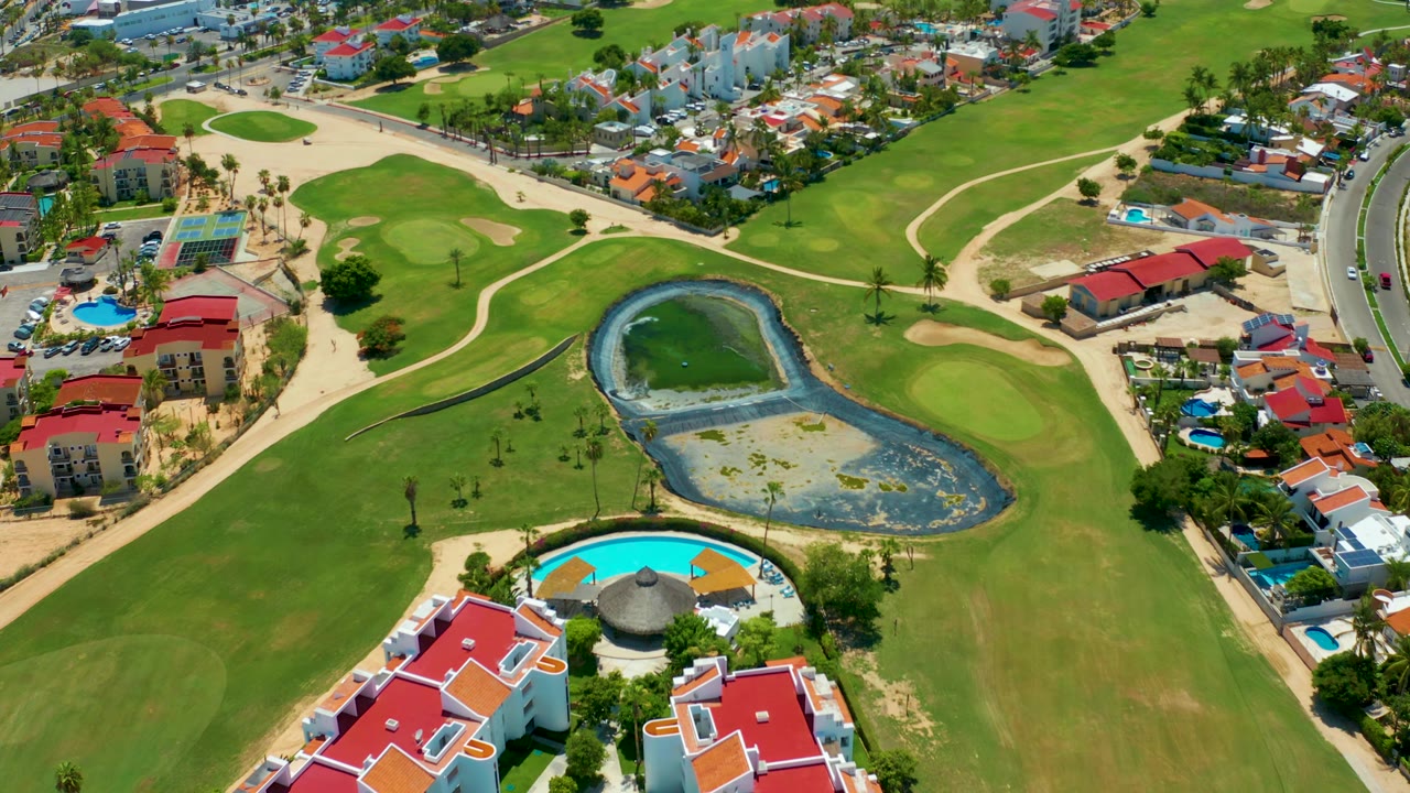 Drone-style aerial view over San Jose Del Cabo with the pond slightly lower in frame, fairways wrapping around it, red-roofed homes nearby, and no visible people.
