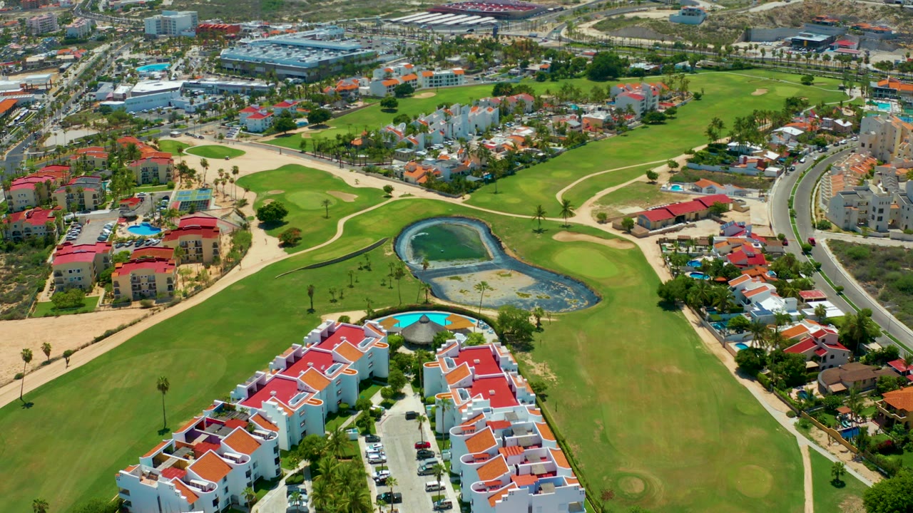 Opening aerial frame over San Jose Del Cabo residences with red roofs in the foreground, green fairways, a small pond, curved roads, and no visible people.