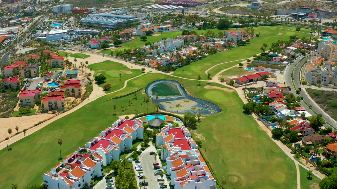Aerial view across fairways, red-roofed homes, pond, and nearby roads.