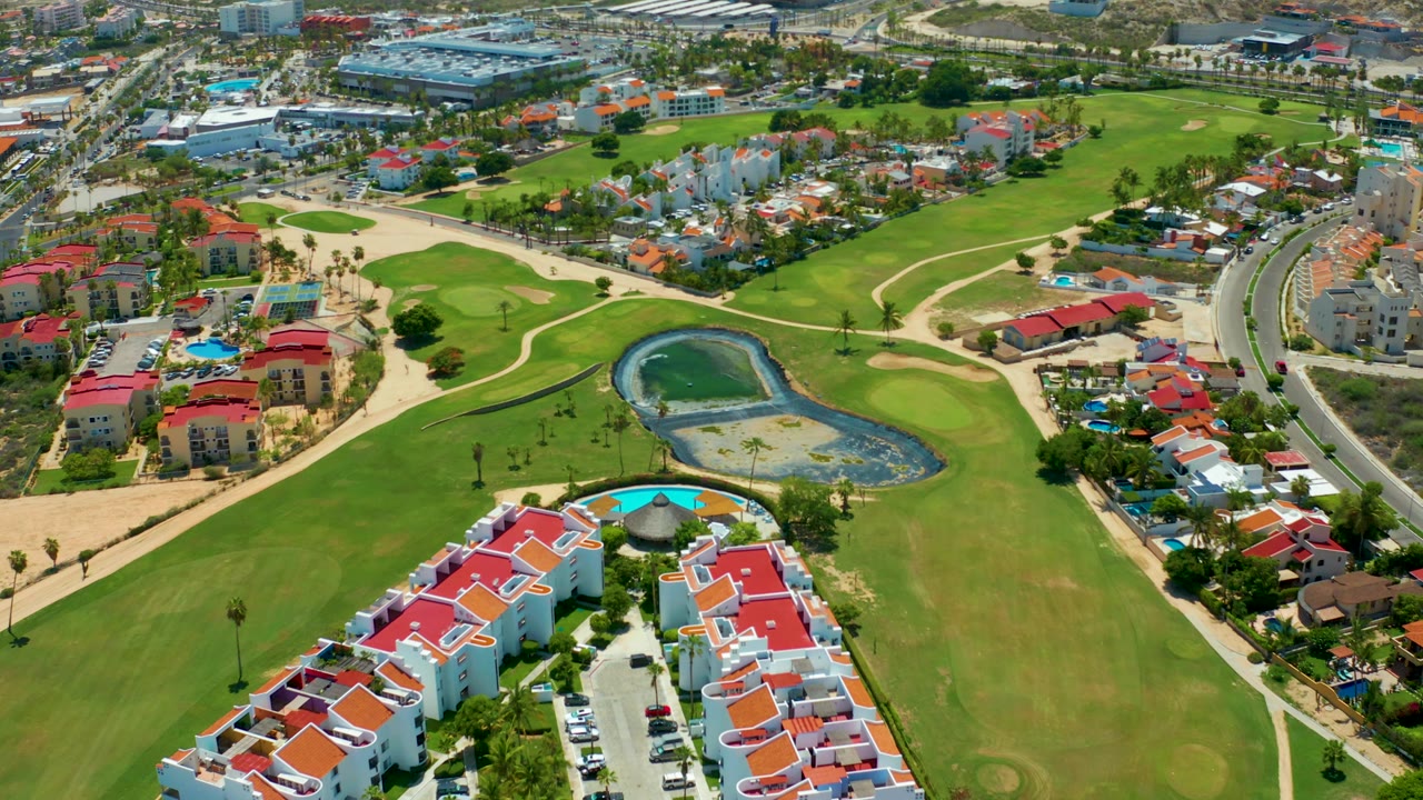Third frame from the elevated San Jose Del Cabo view with the pond on the right side of the fairway, red-roofed homes below, a curved road at the far right, and no visible people.