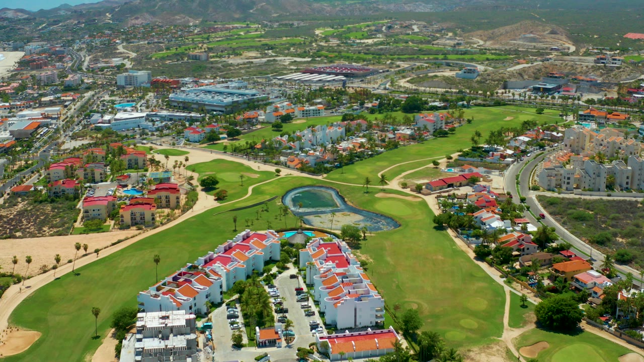 Opening aerial frame over San Jose Del Cabo residences with red roofs, a parking lot, golf fairways, a small pond, curving roads, distant hills, and no visible people.