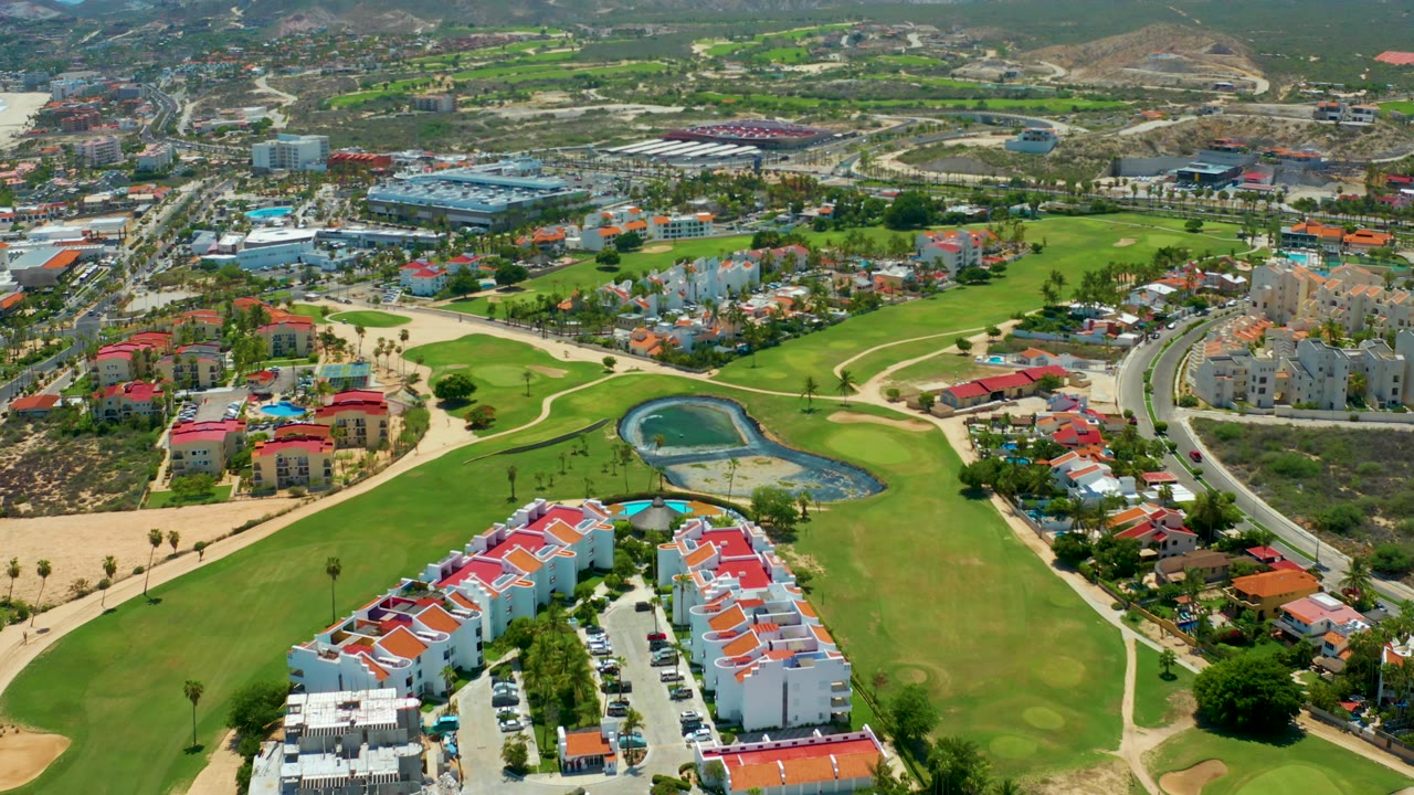 Aerial frame over San Jose Del Cabo with the red-roofed residences lower in frame, a central pond, fairways, road curves, hillside neighborhoods, and no visible people.