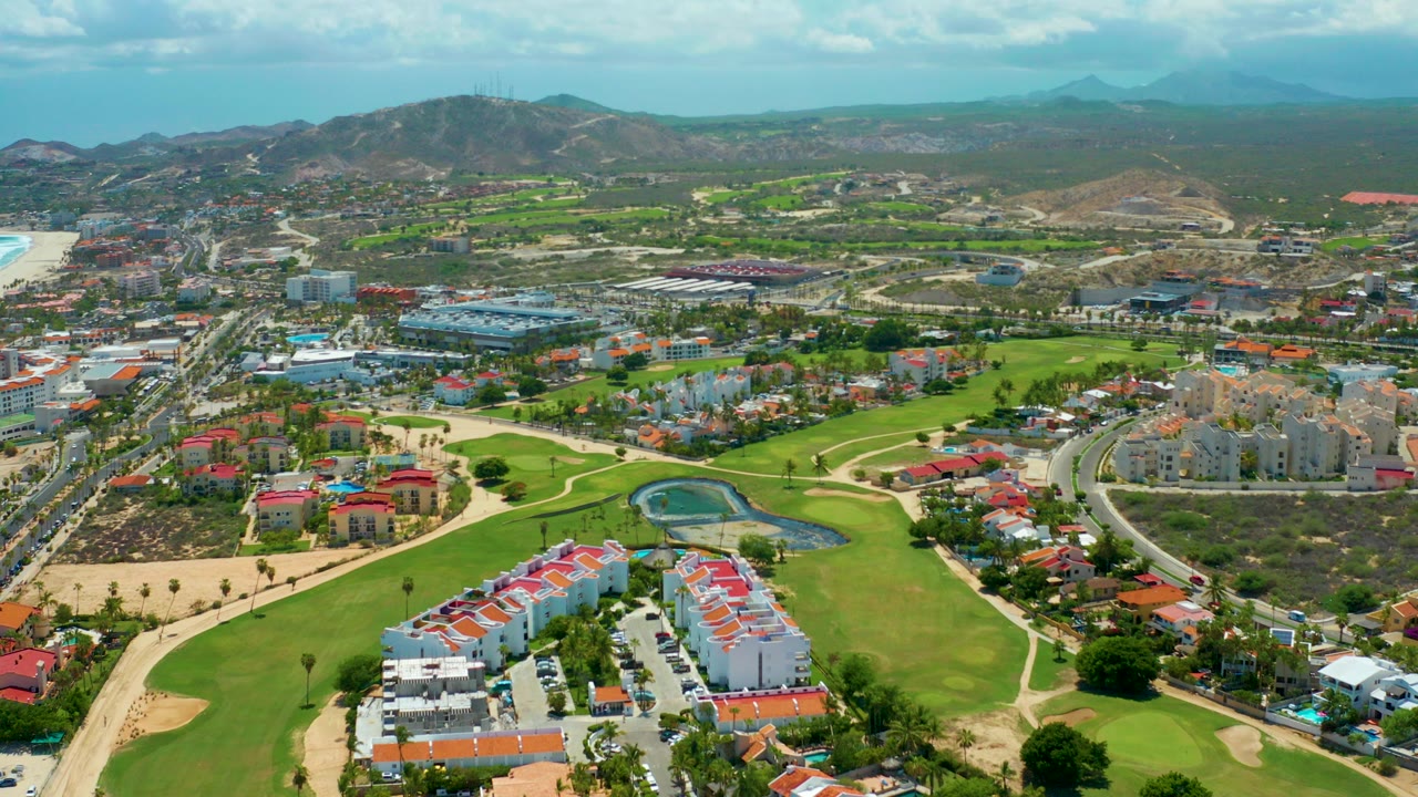 Opening aerial frame over San Jose Del Cabo golf fairways and residential roofs, with a small pond near center right, roads through the midground, mountains on the horizon, and no visible people.