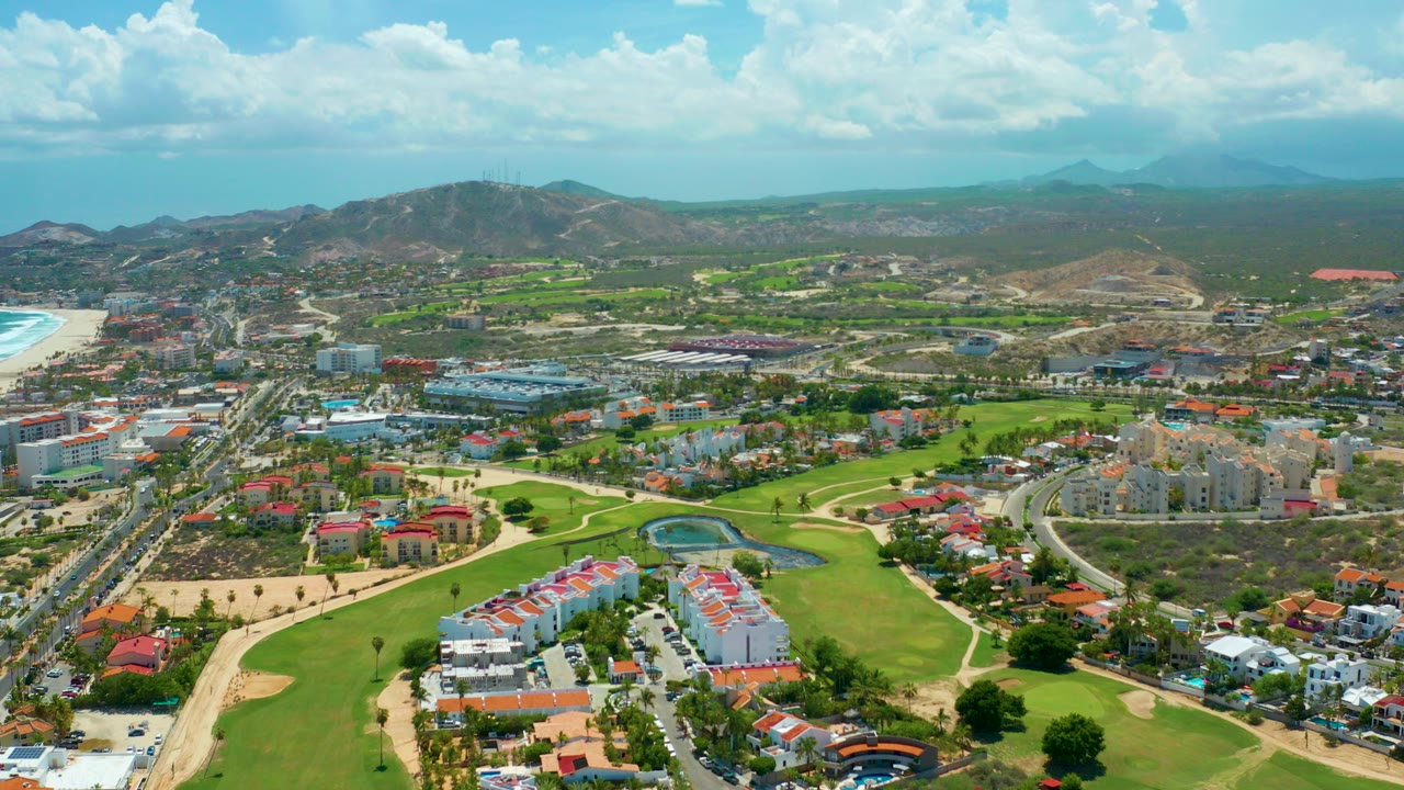 Opening aerial frame over San Jose del Cabo with bright golf greens, low-rise buildings, roads, a beach at left, distant hills and mountains, and no visible people.