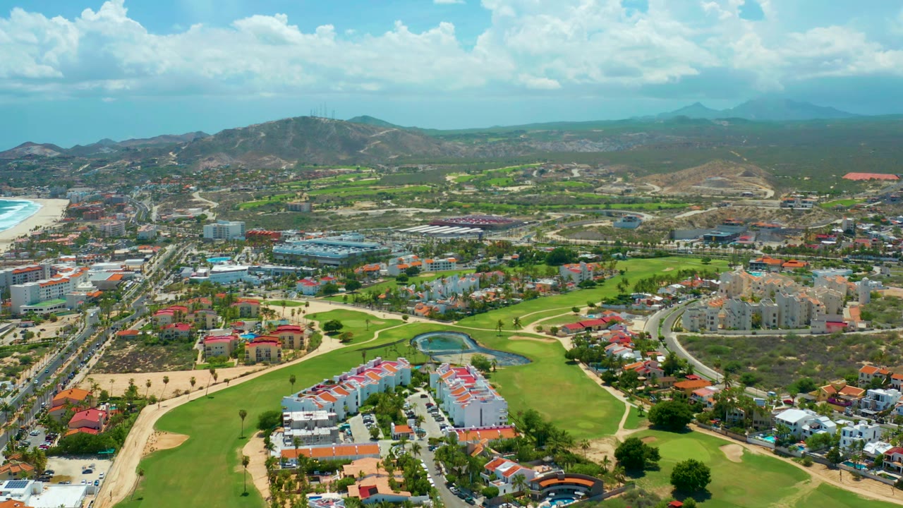 San Jose del Cabo aerial view one second in, with the road corridor and clustered buildings more centered between green fairways and dry hills, with no visible people.