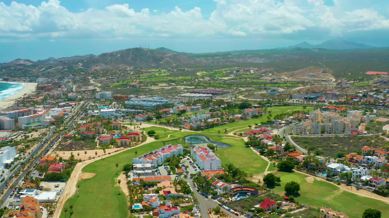 Opening frame over San Jose del Cabo showing green golf fairways, residential buildings, roads, a beach and turquoise ocean at left, dry hills and distant mountains, with no visible people.