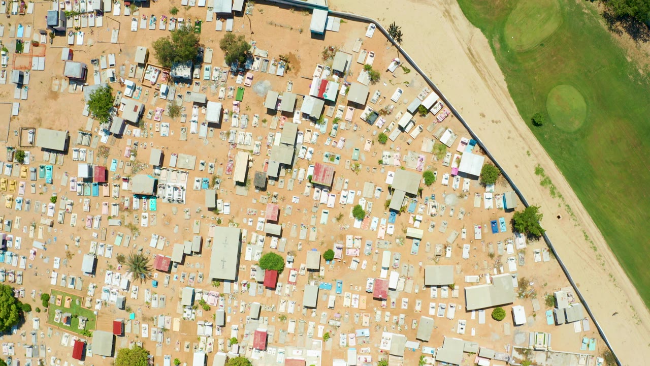 Top-down aerial frame in San Jose del Cabo showing dense small structures on sandy ground next to a green field edge, with no visible people.