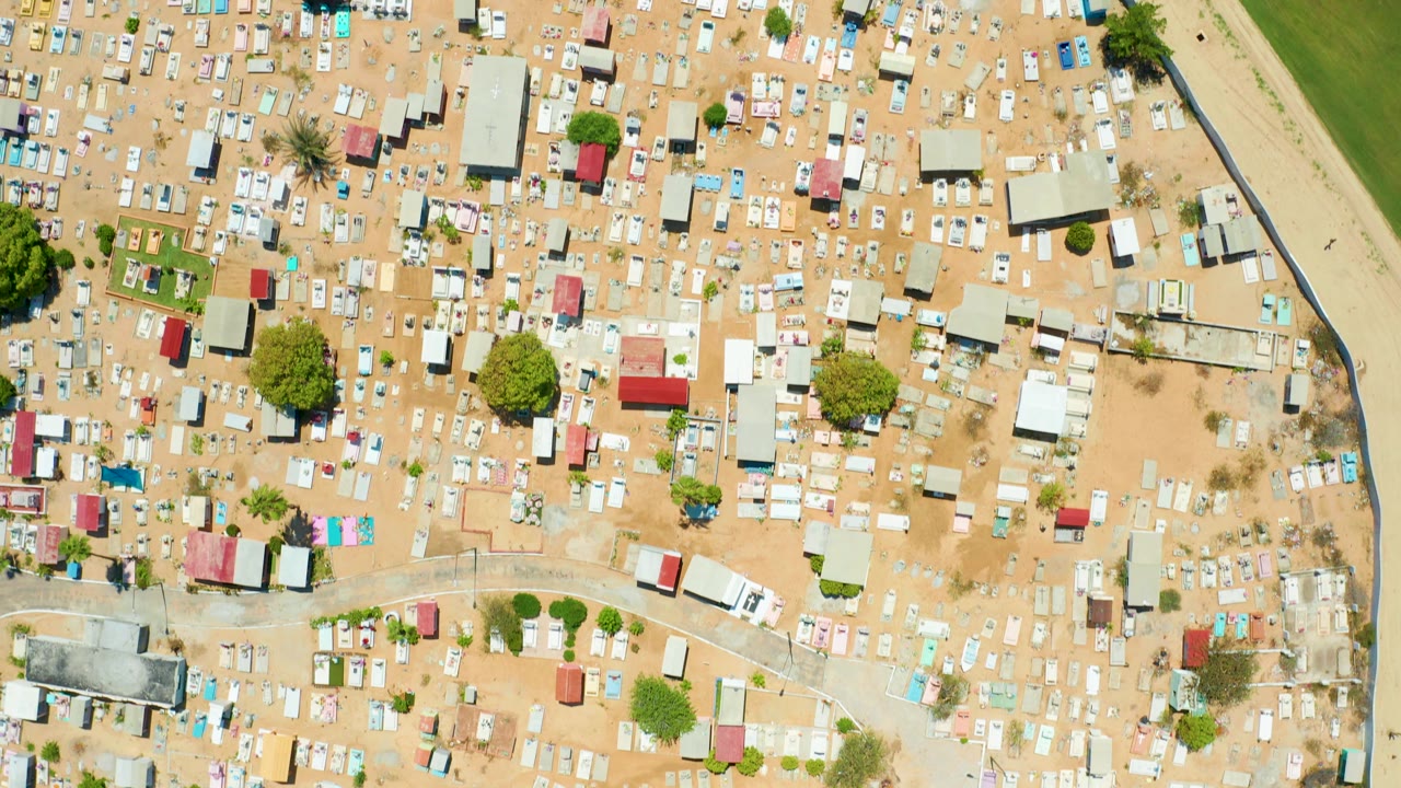High drone frame over San Jose del Cabo with clustered informal structures, sandy open patches, scattered trees, a road, green ground along the right edge, and no visible people.