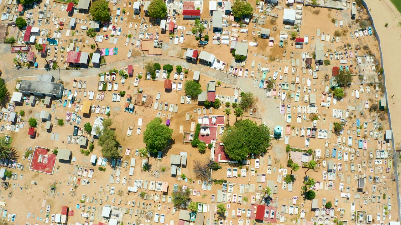 Overhead frame of San Jose Del Cabo showing tightly packed pale grave markers, sandy ground, scattered trees, small roofed structures, and no visible people.