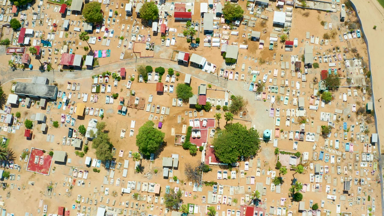 Near-vertical San Jose Del Cabo frame with dense grave plots to the right, scattered small buildings and trees through the center, sandy open ground, and no visible people.