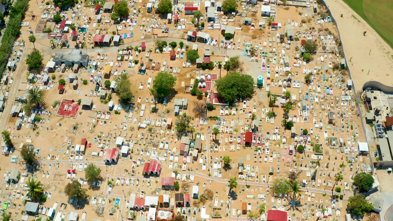 High overhead frame of a temporary campsite in San Jose Del Cabo, Baja California Sur, with dense small tents, sandy paths, green trees, and no visible people.
