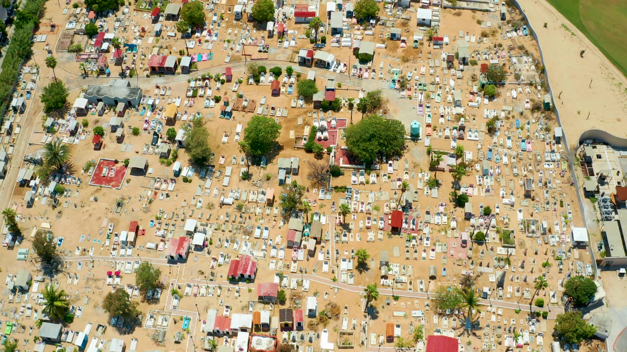 Drone-style overhead view in San Jose Del Cabo showing rows of light tents, dry paths, red-roofed structures, trees, and an edge road or channel with no visible people.