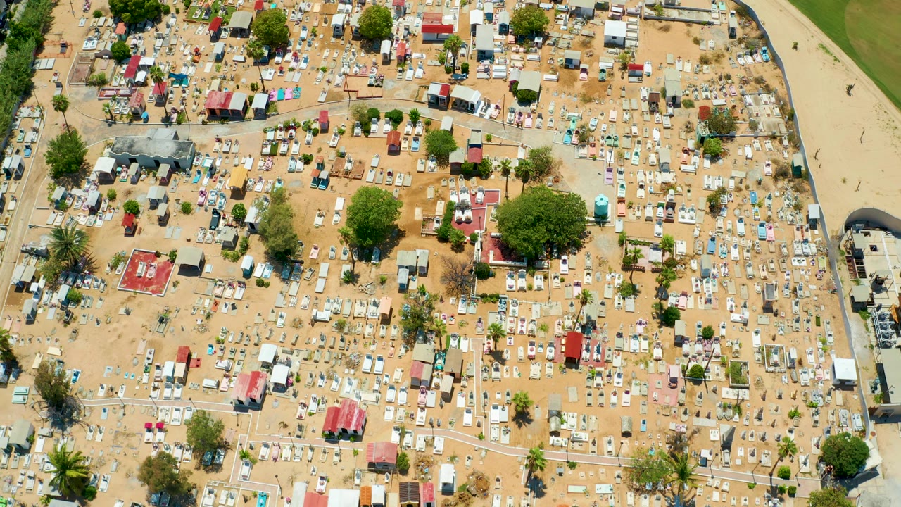 Top-down San Jose Del Cabo campsite frame with red, blue, and white tent roofs, scattered trees, organized footpaths, and no visible people.