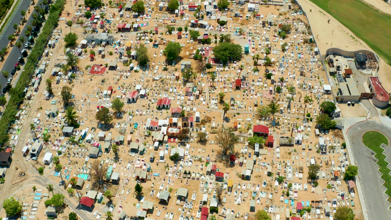 Top-down aerial frame over a sandy RV storage and mobile home area in San Jose Del Cabo, with bordering roads, scattered trees, red-roofed structures, and no visible people.
