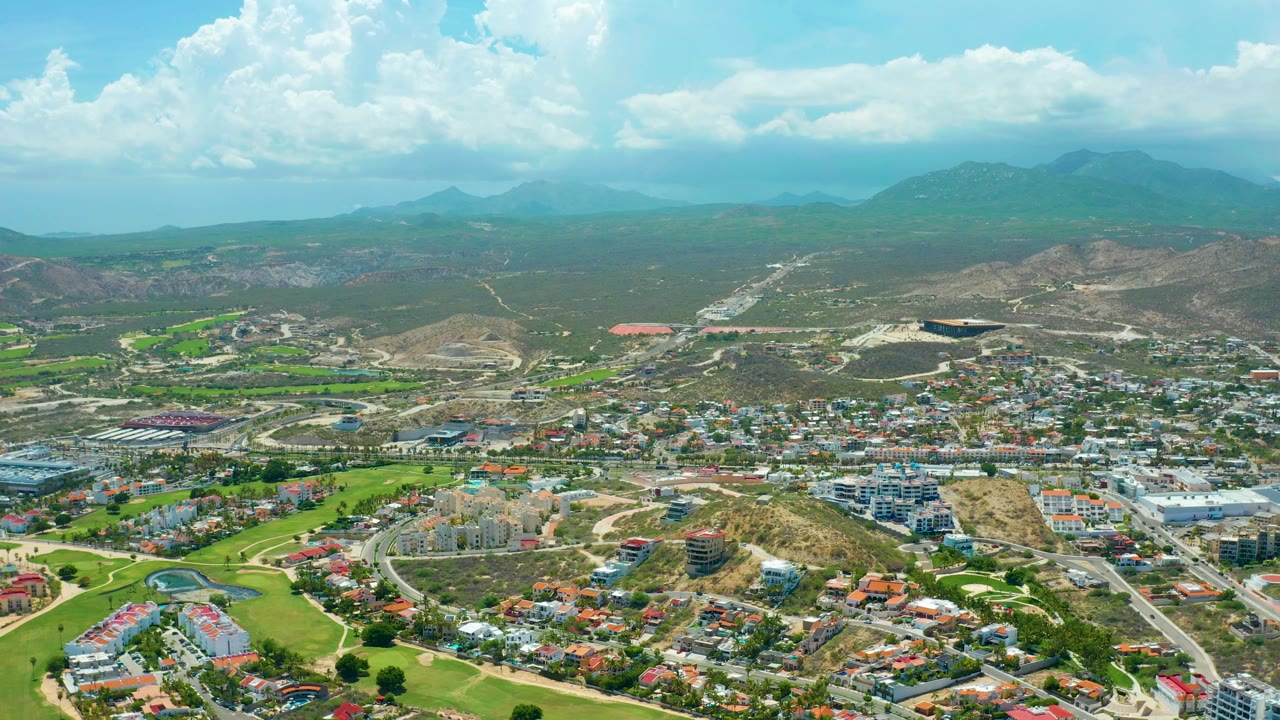 Wide aerial view over San Jose del Cabo with golf course greens and homes in the foreground, hills on the horizon, large white clouds overhead, and no visible people.