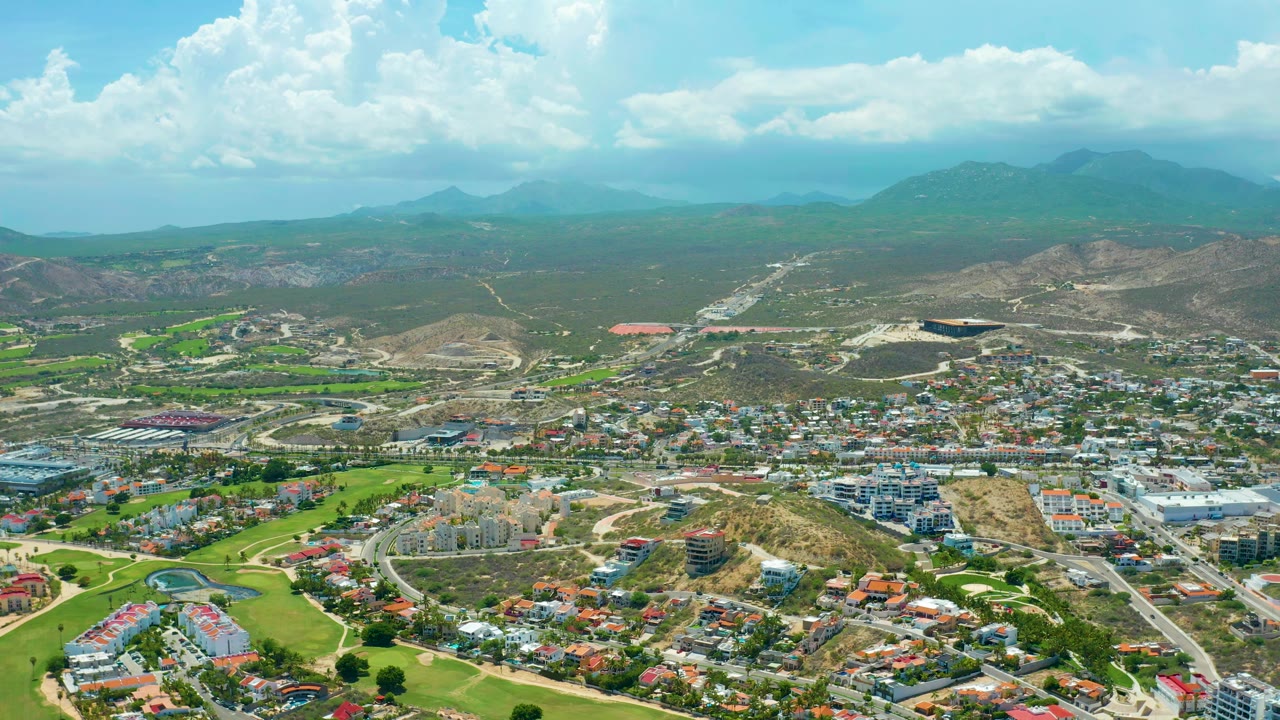San Jose del Cabo appears from a high aerial angle with bright green fairways, dense residential blocks, dry hills beyond the town, and no visible people.