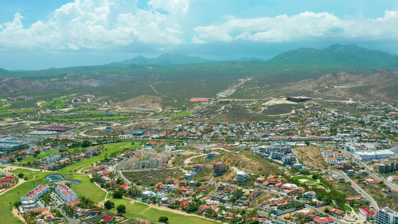 High aerial frame over San Jose del Cabo showing clustered homes, curved roads, green fairways, pale hillside construction areas, distant mountains, and no visible people.