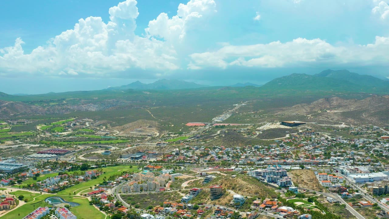 Wide aerial view over San Jose Del Cabo with green fields and buildings below, dry hills and distant mountains beyond, bright cloud cover overhead, and no visible people.