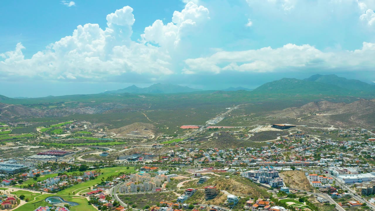 Opening frame of San Jose del Cabo from above, with town blocks and green fields below dry hills, distant mountains, large white clouds, and no visible people.