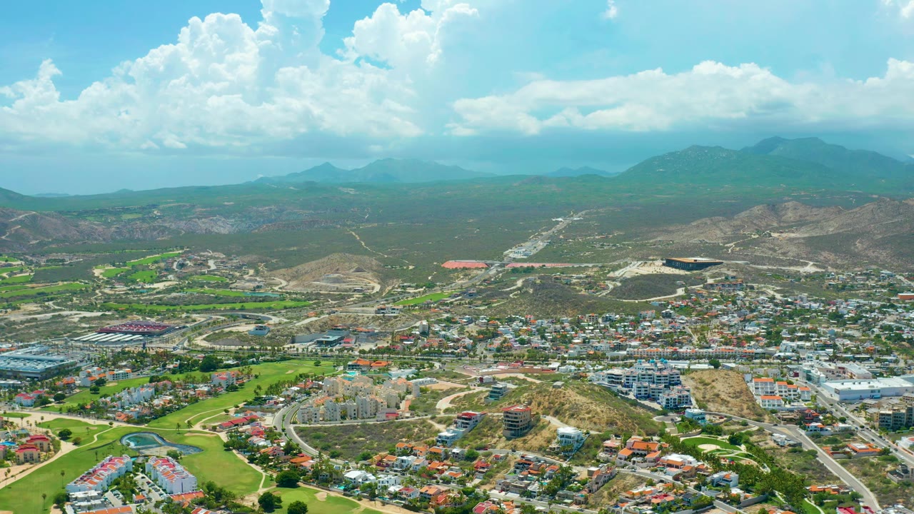 San Jose del Cabo aerial view with clustered buildings in the lower frame, dry hills in the midground, distant mountains under shifting clouds, and no visible people.
